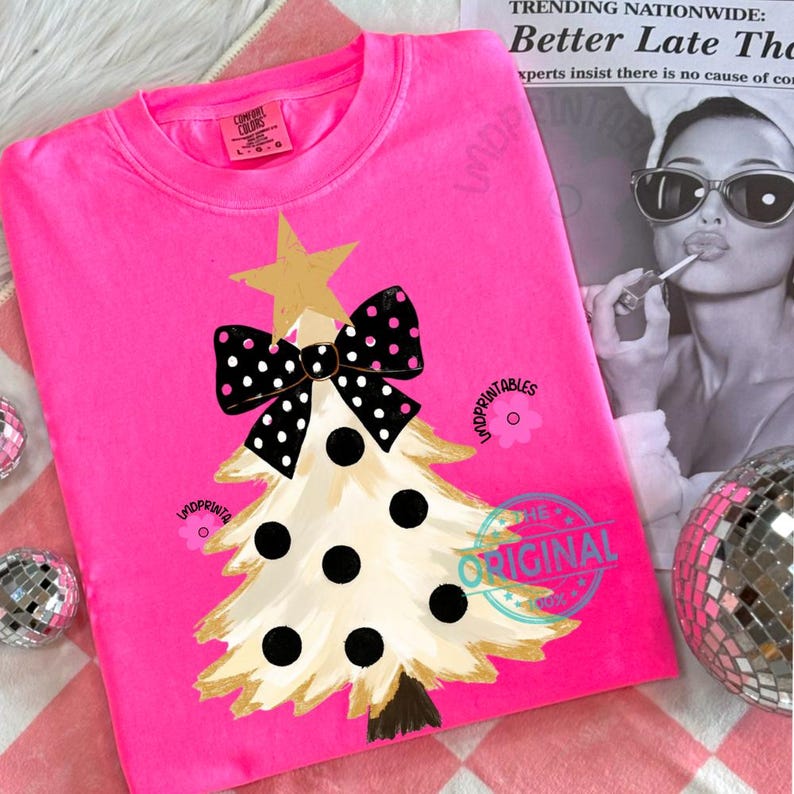 May include: A bright pink sweatshirt featuring a Christmas tree design. The tree is cream-colored with black polka dots and a gold star and bow. The text "THE ORIGINAL" is printed on the shirt.