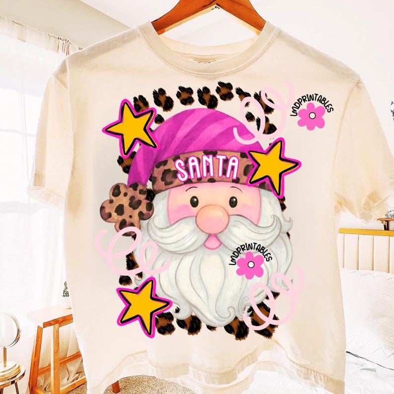 Op de afbeelding: Cr&egrave;mekleurig T-shirt met een afbeelding van de kerstman. De kerstman draagt een roze en luipaardprint hoed met het woord "SANTA". Het ontwerp bevat gele sterren, roze bloemen en luipaardprint accenten, wat een feestelijk vakantiethema cre&euml;ert.