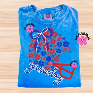 May include: A folded, light blue t-shirt with a floral football helmet design in red and blue. The word "gameday" is written in cursive. The shirt has a Comfort Colors tag.
