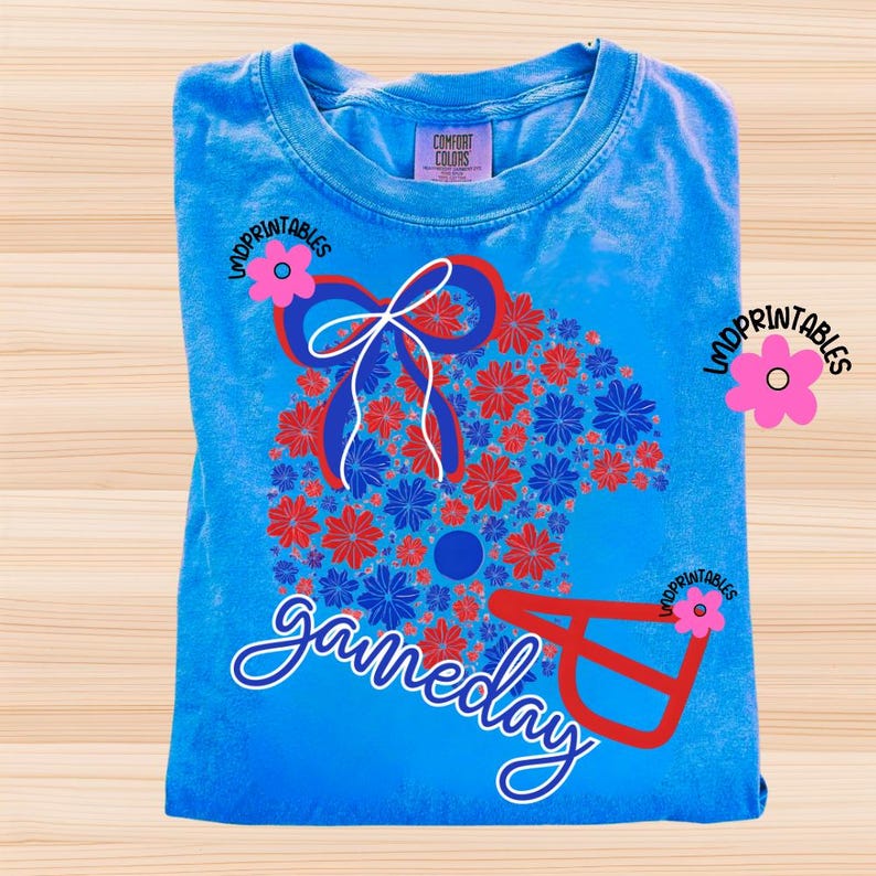 May include: A folded, light blue t-shirt with a floral football helmet design in red and blue. The word "gameday" is written in cursive. The shirt has a Comfort Colors tag.