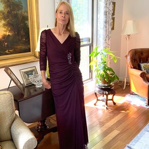May include: A woman wearing a long, dark purple, formal dress with a v-neck and a jeweled brooch. The dress has a ruched design and is made of a sheer fabric.
