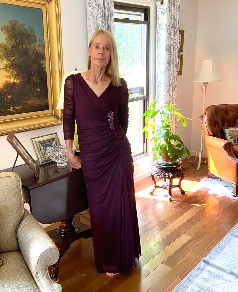 May include: A woman wearing a long, dark purple, formal dress with a v-neck and a jeweled brooch. The dress has a ruched design and is made of a sheer fabric.
