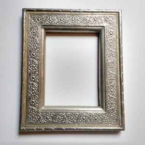 May include: A silver picture frame with an ornate design. The frame has a raised border with a floral pattern.