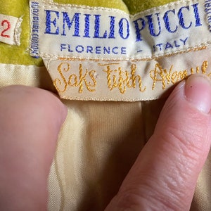 Op de afbeelding: Een geel fluwelen kledingstuk met een wit label waarop "Emilio Pucci, Florence - Itali&euml;" staat en een kleiner wit label waarop "Saks Fifth Avenue" staat.