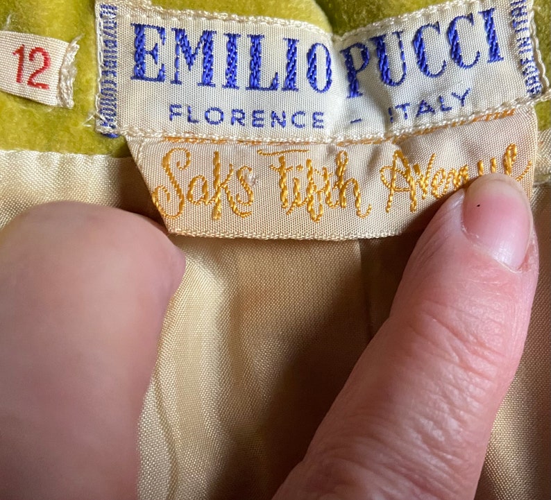 Op de afbeelding: Een geel fluwelen kledingstuk met een wit label waarop "Emilio Pucci, Florence - Itali&euml;" staat en een kleiner wit label waarop "Saks Fifth Avenue" staat.