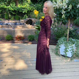 May include: A woman wearing a long, burgundy, ruffled dress with a beaded bodice. She is standing on a wooden deck with a small pond and a white planter with green vines in the background.