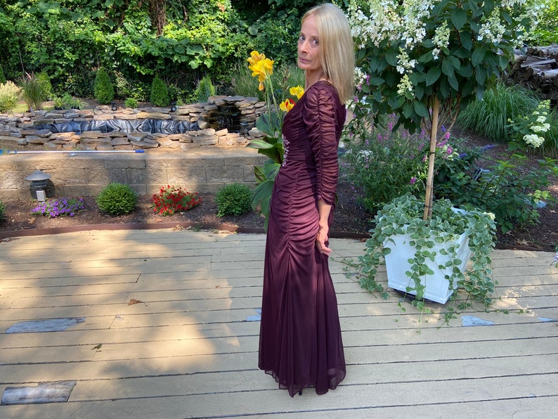 May include: A woman wearing a long, burgundy, ruffled dress with a beaded bodice. She is standing on a wooden deck with a small pond and a white planter with green vines in the background.
