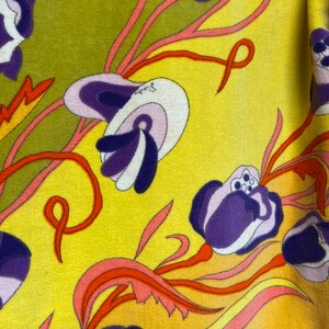 Op de afbeelding: Een gele stof met een bloemenpatroon. De bloemen zijn paars, roze en wit met oranje en rode stelen. De achtergrond is geel.