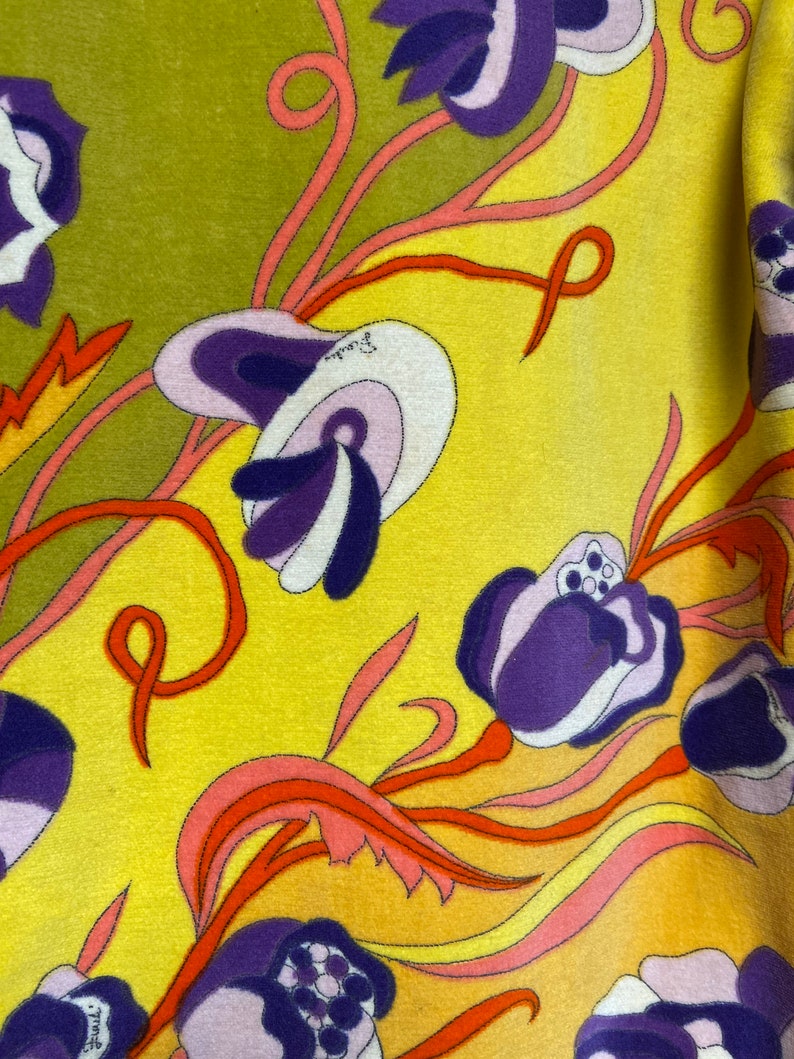 Op de afbeelding: Een gele stof met een bloemenpatroon. De bloemen zijn paars, roze en wit met oranje en rode stelen. De achtergrond is geel.