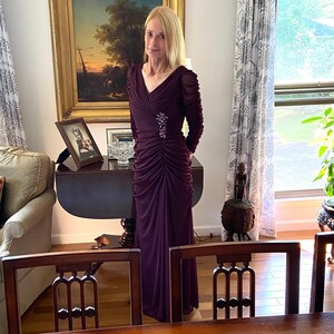 May include: A woman wearing a long, dark purple, ruched dress with a small, sparkly brooch. She is standing in a room with a wooden table and chairs.