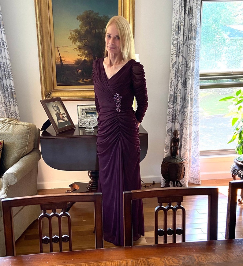 May include: A woman wearing a long, dark purple, ruched dress with a small, sparkly brooch. She is standing in a room with a wooden table and chairs.