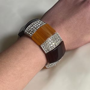 May include: A multi-colored bangle bracelet with a silver metal base and clear rhinestones. The bracelet has three sections of colored resin: orange, brown, and black.