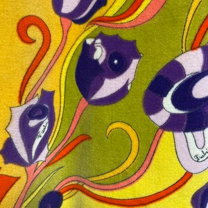 Op de afbeelding: Een gele stof met een kleurrijk bloemenpatroon. De bloemen zijn paars, roze en oranje met een groene achtergrond. De stof heeft een textuur en een subtiele glans.