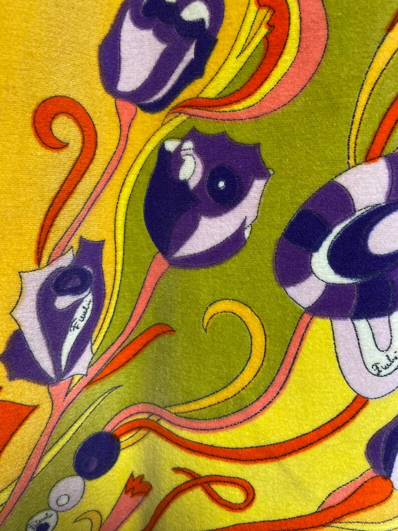 Op de afbeelding: Een gele stof met een kleurrijk bloemenpatroon. De bloemen zijn paars, roze en oranje met een groene achtergrond. De stof heeft een textuur en een subtiele glans.