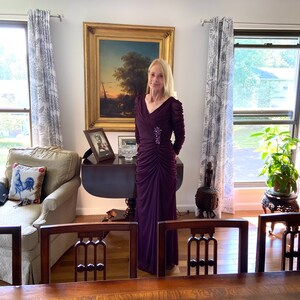 May include: A woman in a long, dark purple dress stands in a room with a painting on the wall, a couch, and a table. She is wearing a small, sparkly brooch on her dress.