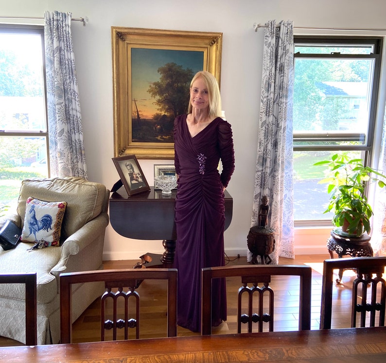 May include: A woman in a long, dark purple dress stands in a room with a painting on the wall, a couch, and a table. She is wearing a small, sparkly brooch on her dress.