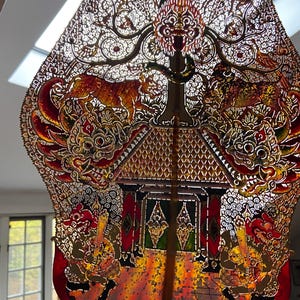 May include: An ornate, intricately carved shadow puppet in warm tones of red, gold, and brown. The puppet features detailed depictions of animals, a tree, and a traditional building, showcasing a rich cultural heritage. The design is symmetrical and illuminated.