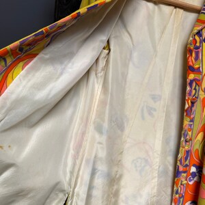 Op de afbeelding: Een vintage jas met een geel, oranje, roze en paars patroon en een witte satijnen voering. De jas heeft een knoopsluiting en een kraag.