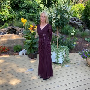 May include: A woman wearing a long, burgundy, sequined dress with a V-neck and long sleeves. She is standing on a wooden deck in a garden setting.