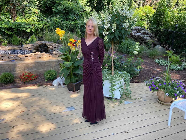 May include: A woman wearing a long, burgundy, sequined dress with a V-neck and long sleeves. She is standing on a wooden deck in a garden setting.