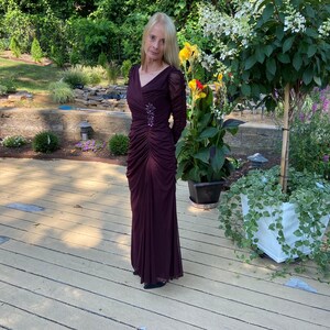 May include: A woman wearing a long, burgundy, formal dress with a beaded embellishment on the bodice. She is standing on a wooden deck with a potted plant and flowers in the background.