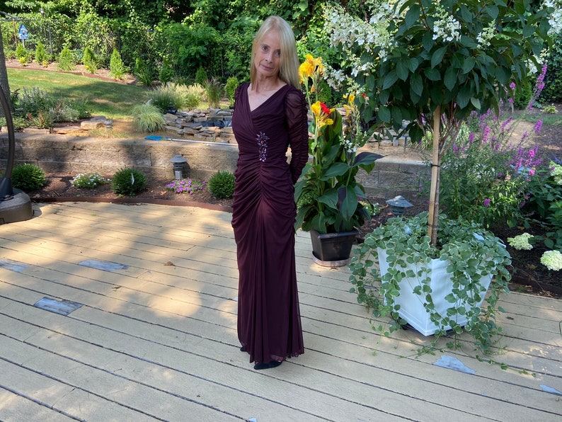 May include: A woman wearing a long, burgundy, formal dress with a beaded embellishment on the bodice. She is standing on a wooden deck with a potted plant and flowers in the background.