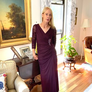 May include: A woman wearing a long, dark purple, formal dress with a beaded embellishment on the bodice. The dress has a V-neckline and long sleeves. She is standing in a room with hardwood floors and a painting on the wall.