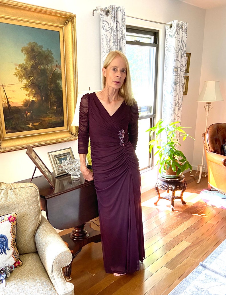 May include: A woman wearing a long, dark purple, formal dress with a beaded embellishment on the bodice. The dress has a V-neckline and long sleeves. She is standing in a room with hardwood floors and a painting on the wall.