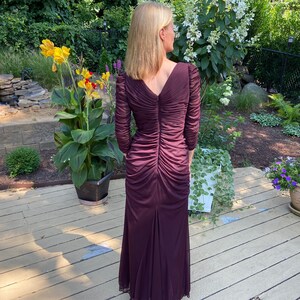 May include: A long, burgundy-colored dress with a V-neckline and long sleeves. The dress has a ruched design and a fitted silhouette.