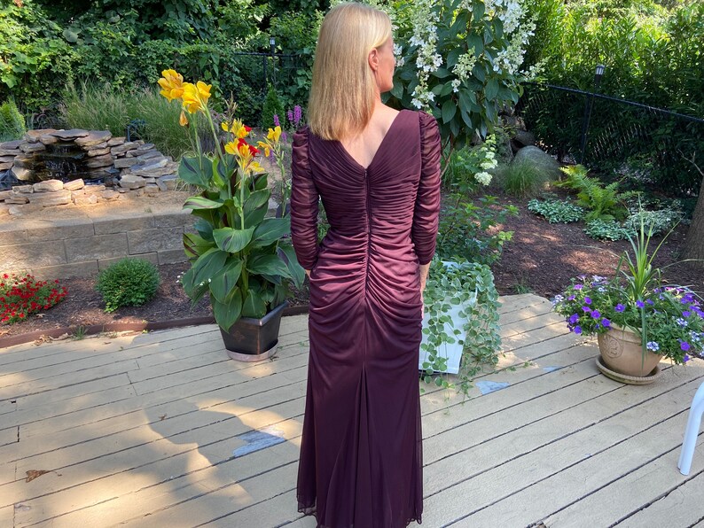 May include: A long, burgundy-colored dress with a V-neckline and long sleeves. The dress has a ruched design and a fitted silhouette.