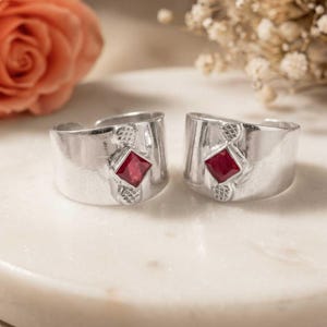 May include: Two wide silver rings featuring a polished finish and a central square-cut red gemstone. Each ring has a textured, cross-hatched detail above and below the stone, with an open-back design that allows for adjustable sizing on the finger.