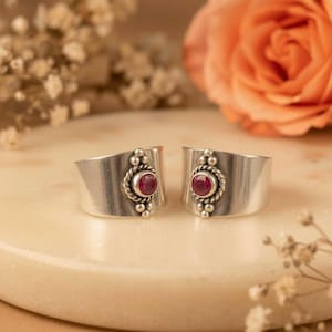 Peut inclure: Deux bagues larges en argent avec une pierre rouge ronde à facettes au centre. Chaque pierre est sertie dans une bordure en corde torsadée décorative et ornée de petites sphères en argent au-dessus et en dessous du sertissage.