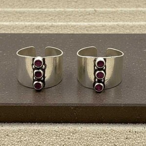 Può includere: Due anelli larghi a fascia aperta realizzati in metallo color argento lucido. Ogni anello presenta una fila verticale di tre piccole pietre circolari rosso scuro incastonate in un canale scuro e incassato al centro della fascia.
