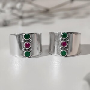 May include: Two wide silver rings, each featuring a vertical row of three bezel-set stones. The center stone is a vibrant magenta-pink, flanked by two deep green stones. The metal has a smooth, polished finish with a simple, clean band design.