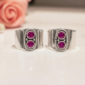 Peut inclure: Deux bagues larges en argent, chacune dotée d'un sertissage ovale vertical central. Ce sertissage contient deux pierres rondes rose foncé disposées verticalement, encadrées par une bordure en corde torsadée et de petites perles d'argent.