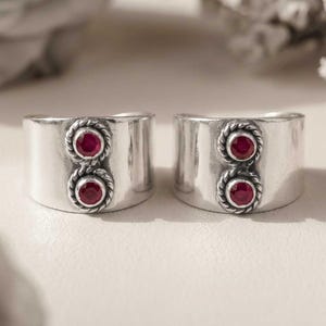 May include: Two wide silver rings, each featuring a vertical pair of round, deep red rubies. Each ruby is set within a decorative, twisted rope-style silver border, contrasting against the smooth, polished surface of the wide metal bands.