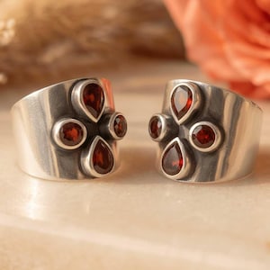 May include: Two wide silver rings, each featuring a cluster of four deep red garnet gemstones. The stones are arranged in a cross pattern, with two round-cut garnets and two pear-shaped garnets set into the smooth, polished silver band surface.