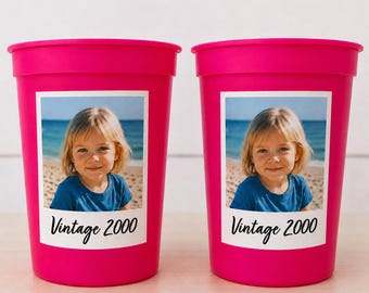 Puede incluir: Dos vasos de plástico rosa brillante. Cada uno tiene un rectángulo blanco con una foto de un niño en la playa. Debajo de la foto está el texto "Vintage 2000" en una fuente manuscrita.