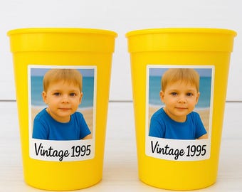 Puede incluir: Dos vasos de plástico amarillos brillantes. Cada vaso tiene una etiqueta blanca con una foto de un niño pequeño con una camisa azul en una playa. La etiqueta también tiene el texto "Vintage 1995" en cursiva.