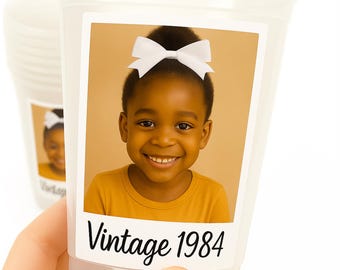Puede incluir: Vaso de plástico transparente con una etiqueta que muestra a un niño sonriente con un lazo blanco y una camiseta amarilla. La etiqueta dice "Vintage 1984" en escritura negra. Otro vaso es visible en el fondo.