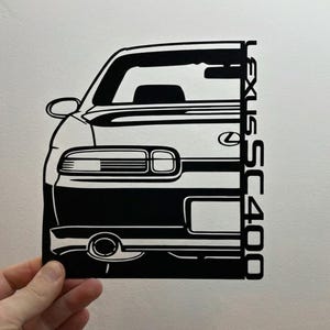 May include: Black and white die-cut of a Lexus SC400 car. The image shows the front and side profile of the vehicle, with details like the headlights, side mirror, and the car's name printed vertically on the side.