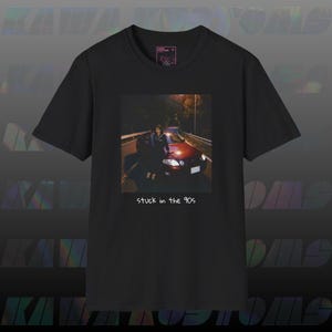 May include: Black t-shirt featuring a photograph of a person standing next to a red car at night. The image has the text "stuck in the 90s" in white at the bottom. The shirt has a crew neck and short sleeves.