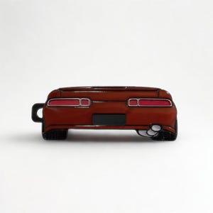 May include: A brown car-shaped pin with red taillights and a black license plate area. The pin has a black hook on the left side and a silver exhaust pipe on the right. The pin is set against a white background.