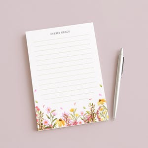 May include: A lined notepad with the text "EVERLY GRACE" at the top, featuring a floral design at the bottom with pink, yellow, and green flowers. A silver pen rests beside the notepad on a light pink surface.