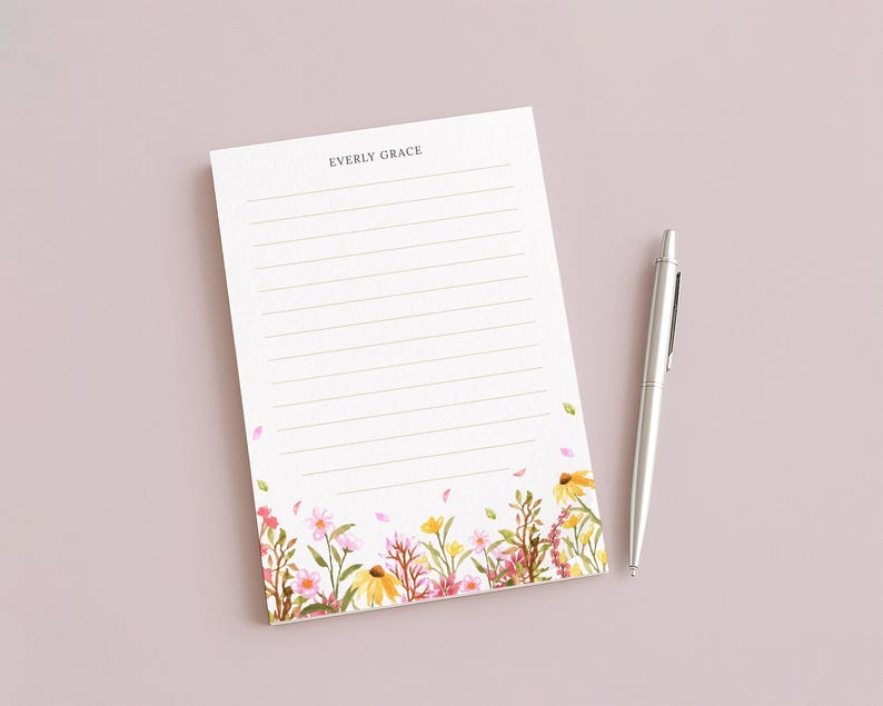 May include: A lined notepad with the text "EVERLY GRACE" at the top, featuring a floral design at the bottom with pink, yellow, and green flowers. A silver pen rests beside the notepad on a light pink surface.