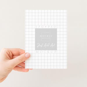 Peut inclure: Une main tenant une carte blanche avec un motif quadrill&eacute;. La carte pr&eacute;sente le texte "JPG &bull; PSD" et "Mockup Days Co" avec "Just Add Art" dans un rectangle gris. Le fond est de couleur blanc cass&eacute;.