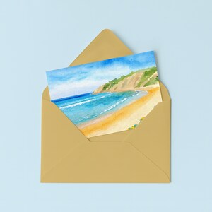 May include: A watercolor painting of a beach scene, featuring a sandy shore, turquoise water, and a green and tan cliffside. The artwork is inside a gold envelope, set against a light blue background.