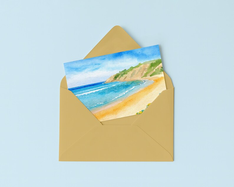 May include: A watercolor painting of a beach scene, featuring a sandy shore, turquoise water, and a green and tan cliffside. The artwork is inside a gold envelope, set against a light blue background.