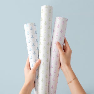 May include: Three rolls of white wrapping paper with a repeating anchor pattern in blue, green, and pink. The rolls are held against a light blue background. The paper is suitable for nautical or coastal themed gifts.