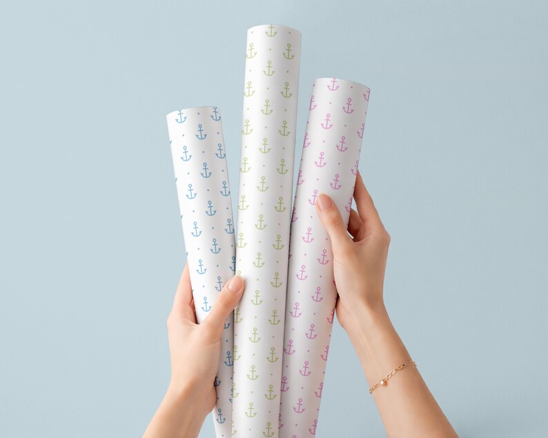 May include: Three rolls of white wrapping paper with a repeating anchor pattern in blue, green, and pink. The rolls are held against a light blue background. The paper is suitable for nautical or coastal themed gifts.